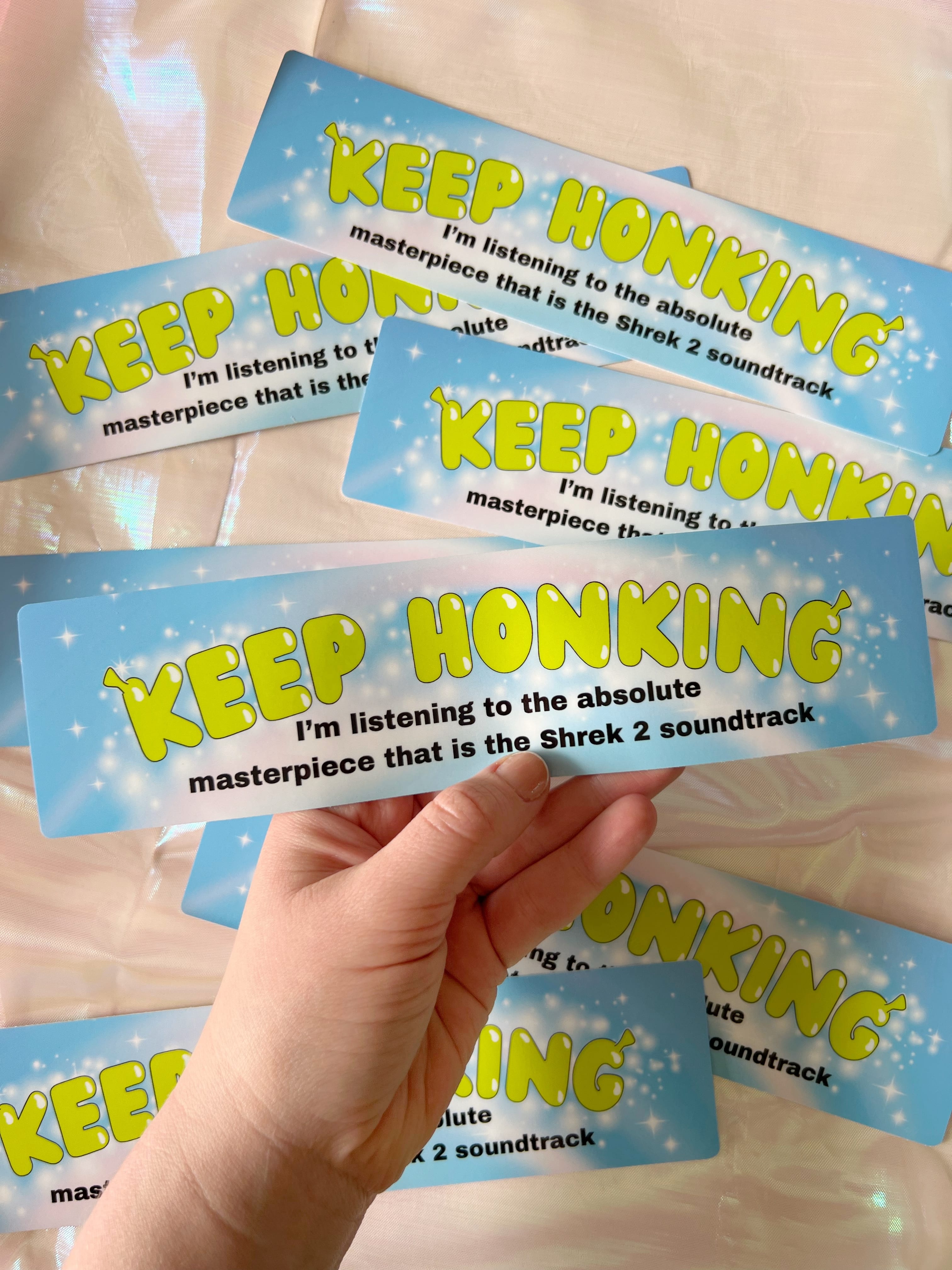 a hand holding a bumper sticker that says "keep honking I'm listening to the absolute masterpiece that is the Shrek 2 soundtrack"