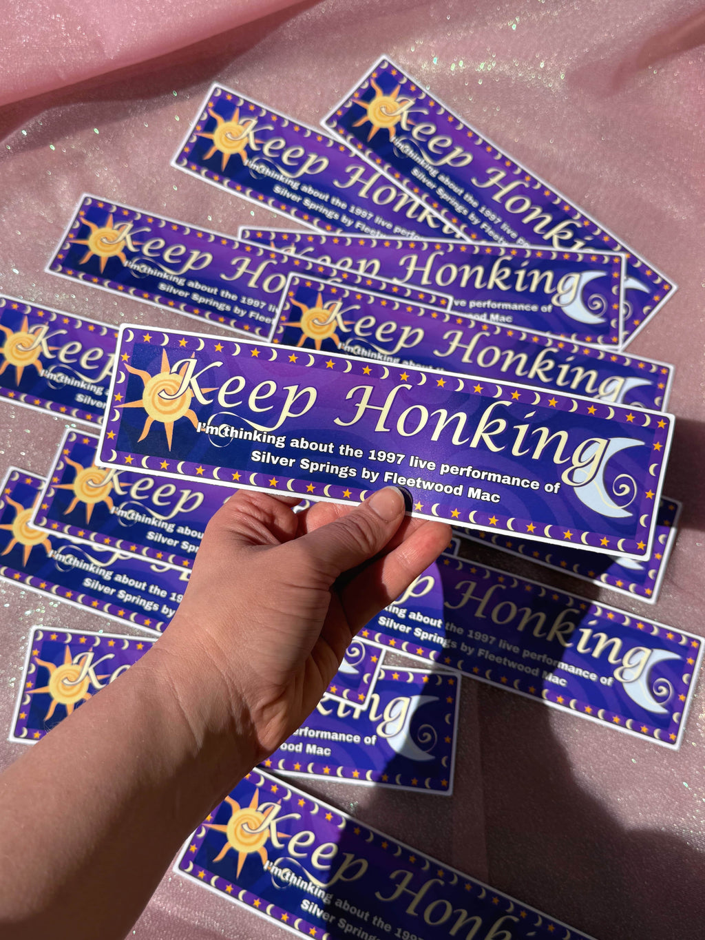 'Keep Honking I'm Thinking about the 1997 live performance of Silver Springs by Fleetwood Mac' bumper stickers with a hand holding one, on a pink surface.