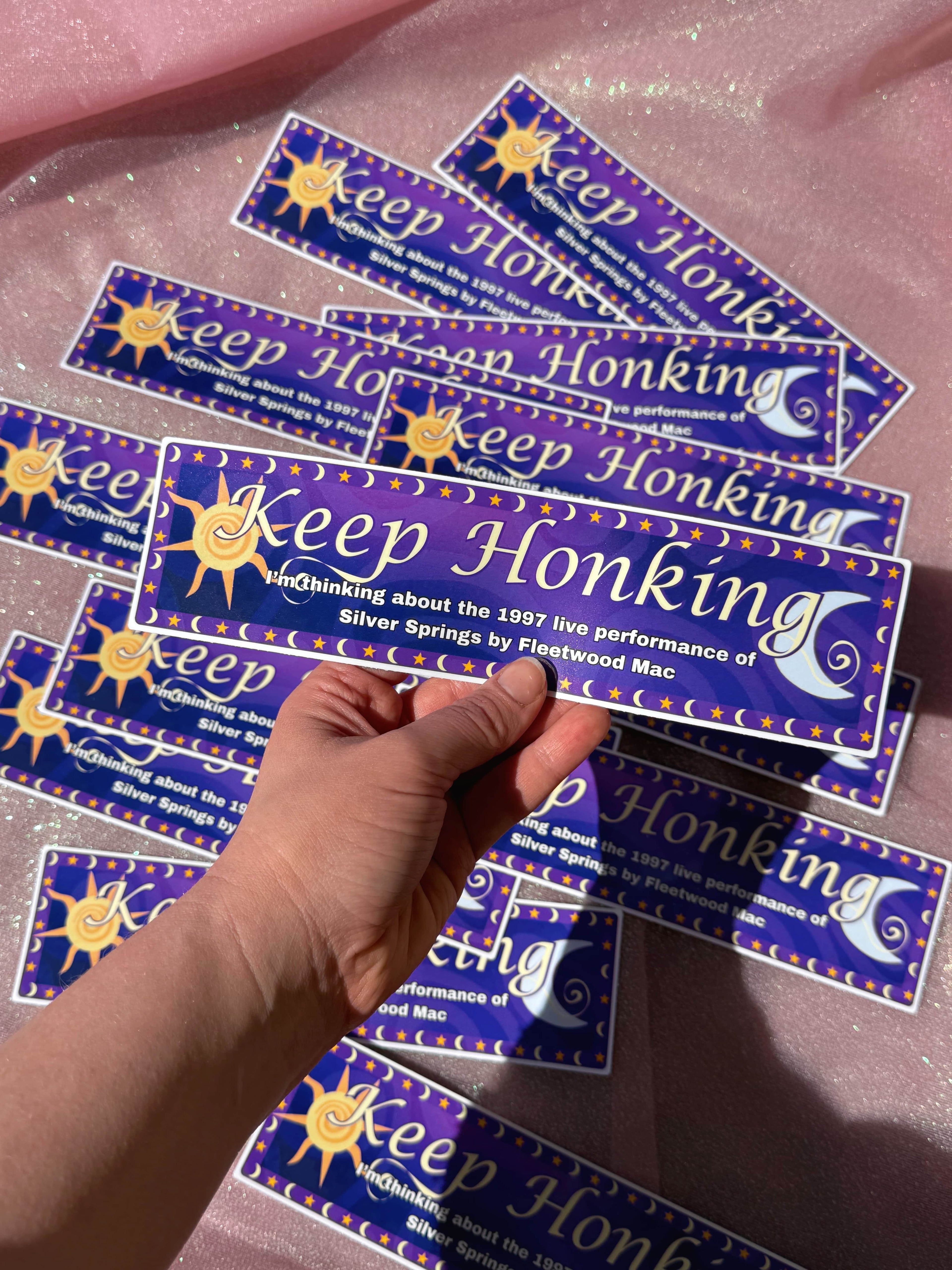 'Keep Honking I'm Thinking about the 1997 live performance of Silver Springs by Fleetwood Mac' bumper stickers with a hand holding one, on a pink surface.