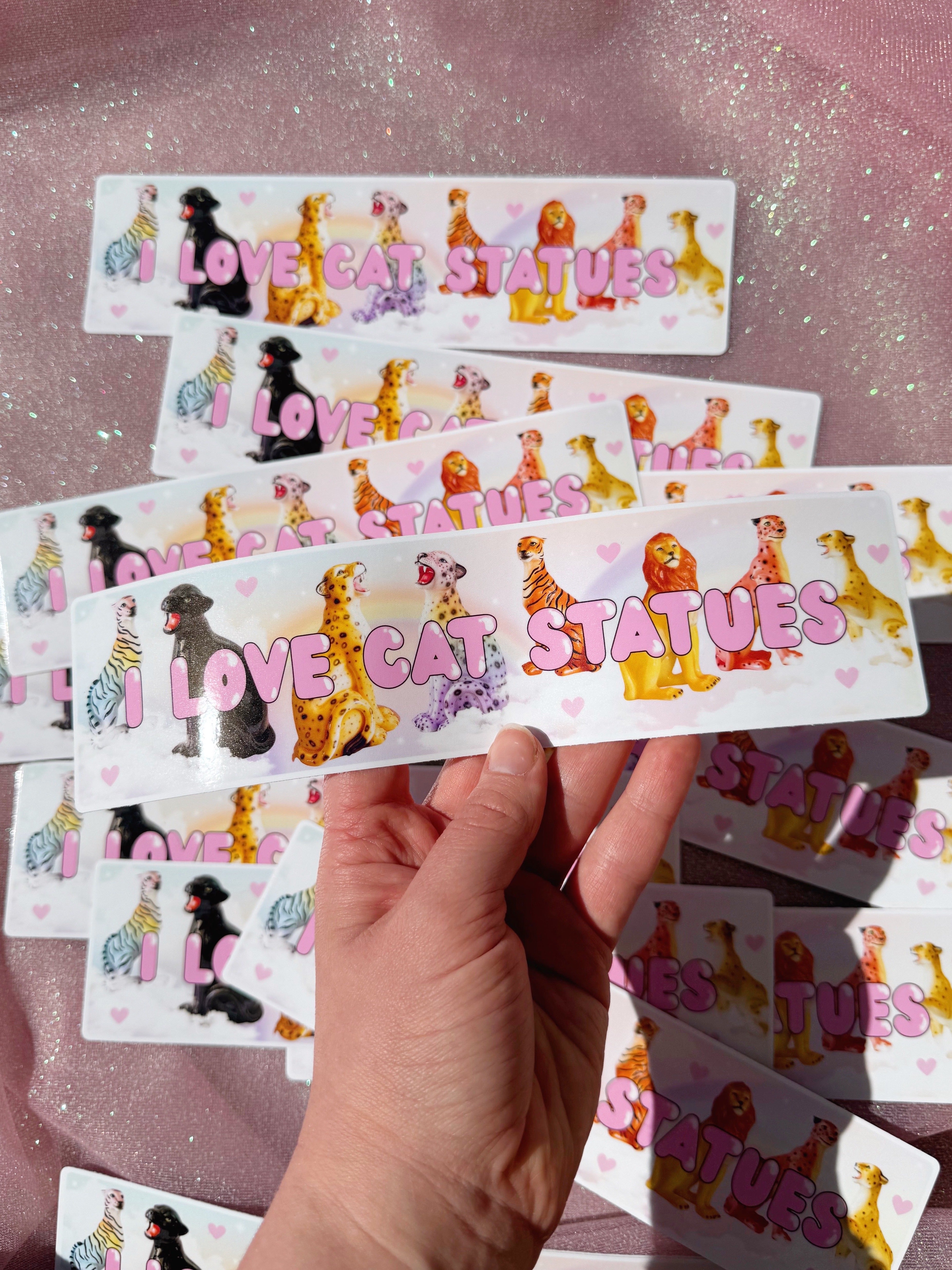 Hand holding a sheet of 'I Love Cat Statues' stickers with various cat illustrations on a pink glittery background.