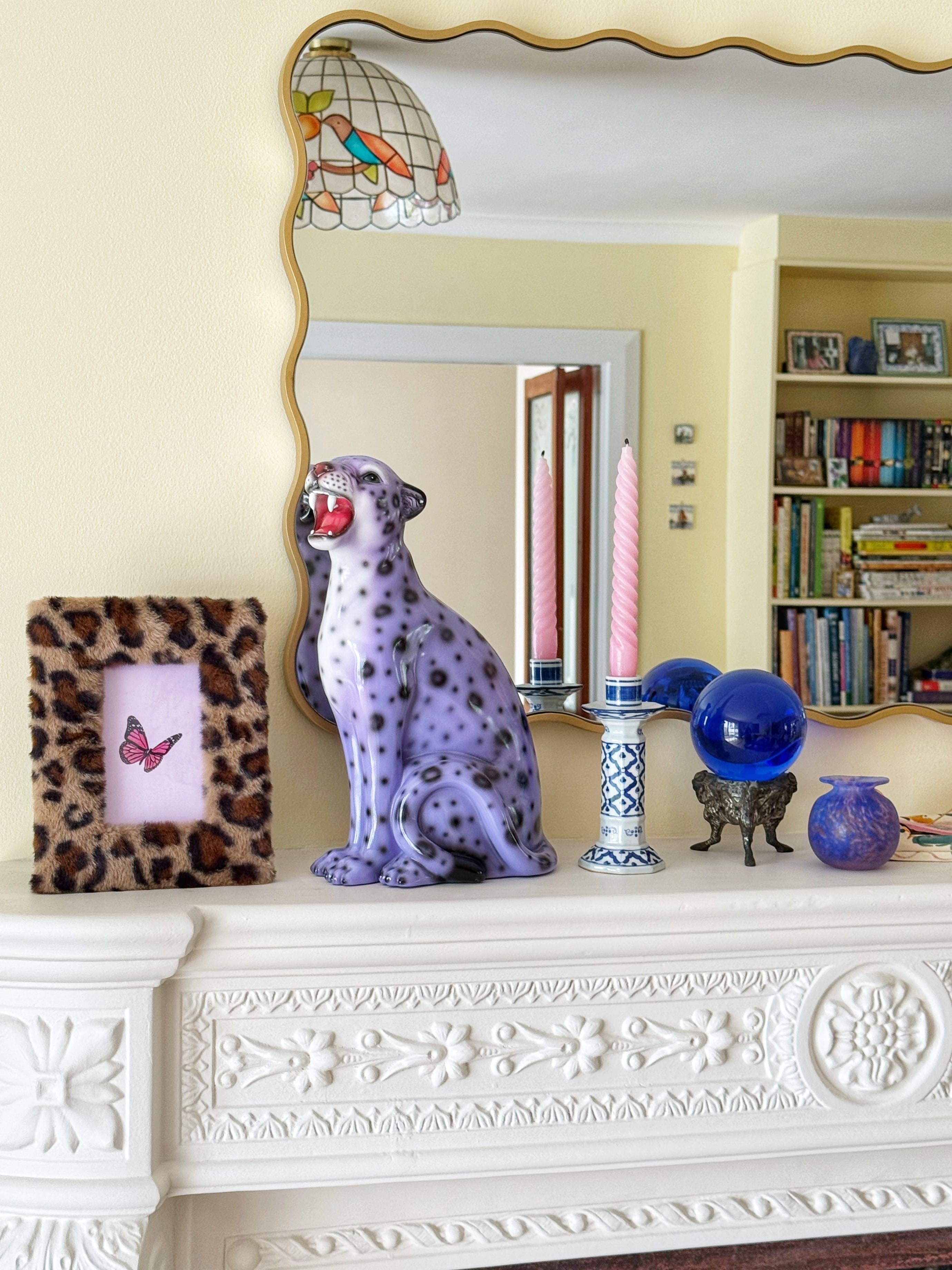 Purple leopard statue and other decorative items on a mantelpiece with a mirror reflecting a bookshelf.