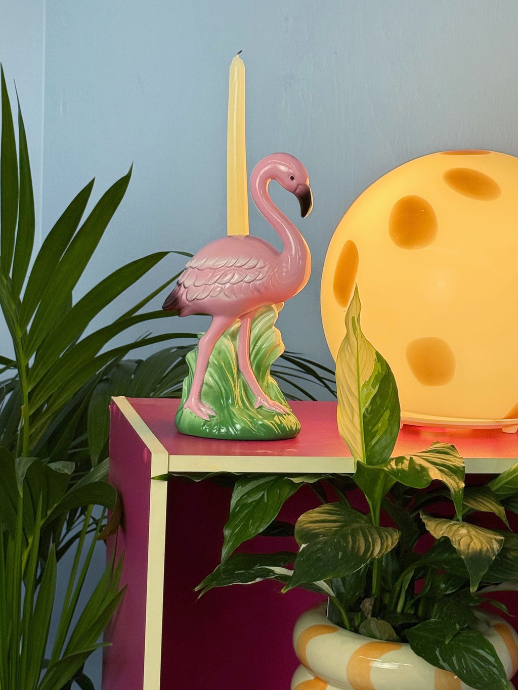 Pink flamingo candle holder on a pink table with a yellow moon lamp in the background.