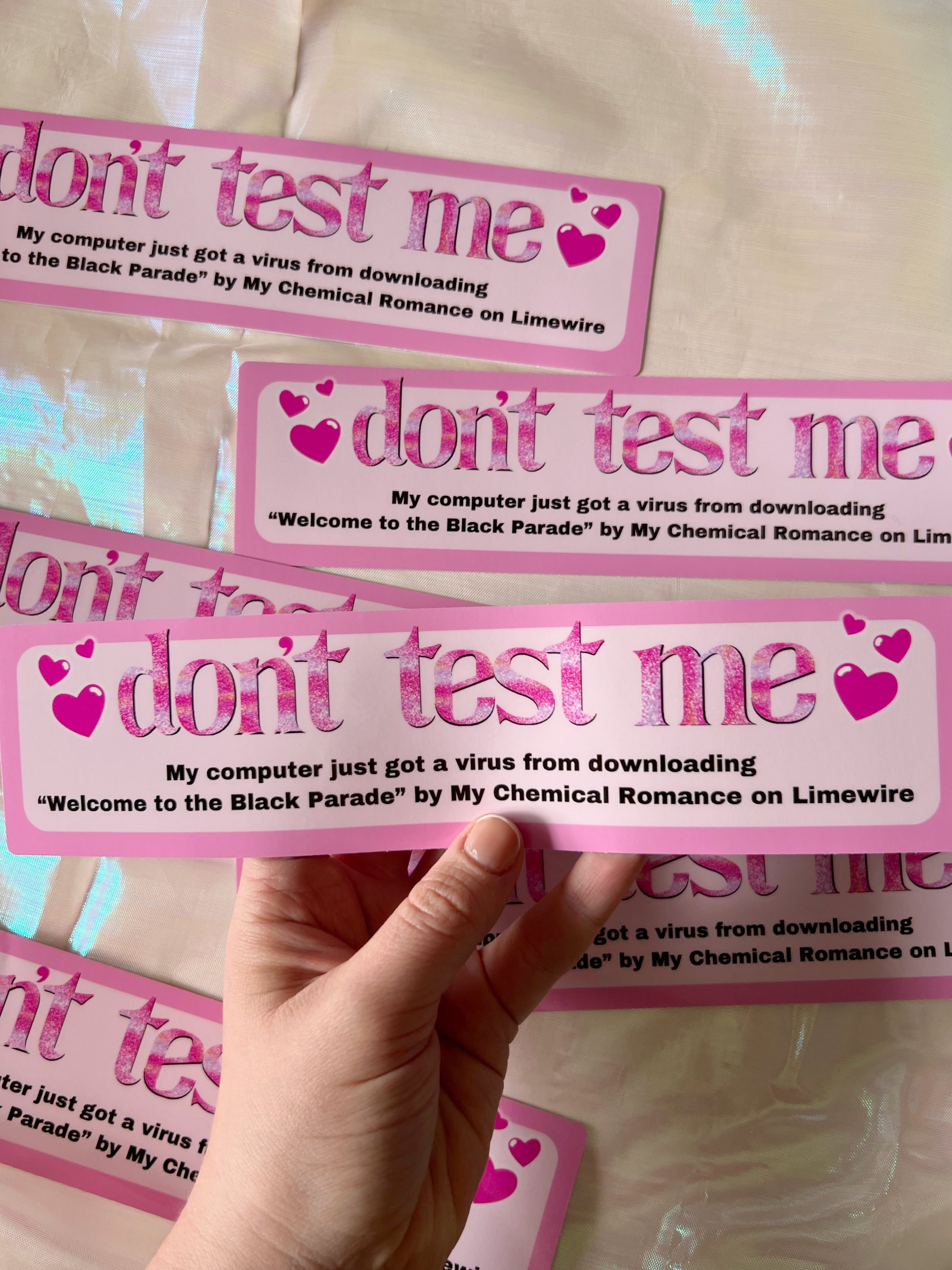 a hand holding a bumper sticker that says "don't test me my computer just got a virus from downloading welcome to the black parade by my chemical romance on limewire"